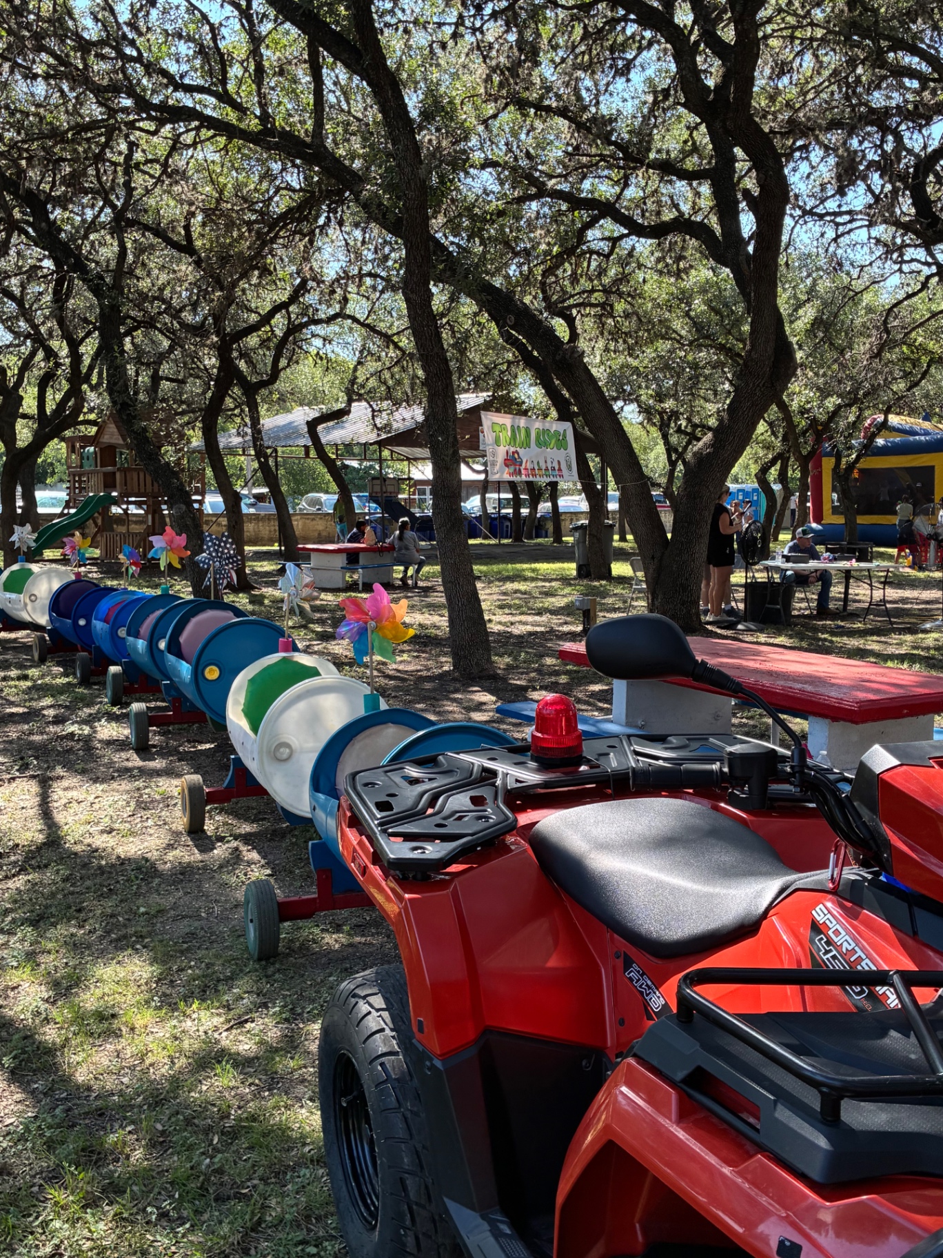The colorful Doodle Train cars parked under shady oak trees at a park event, with a Train Rides banner and bounce house behind them