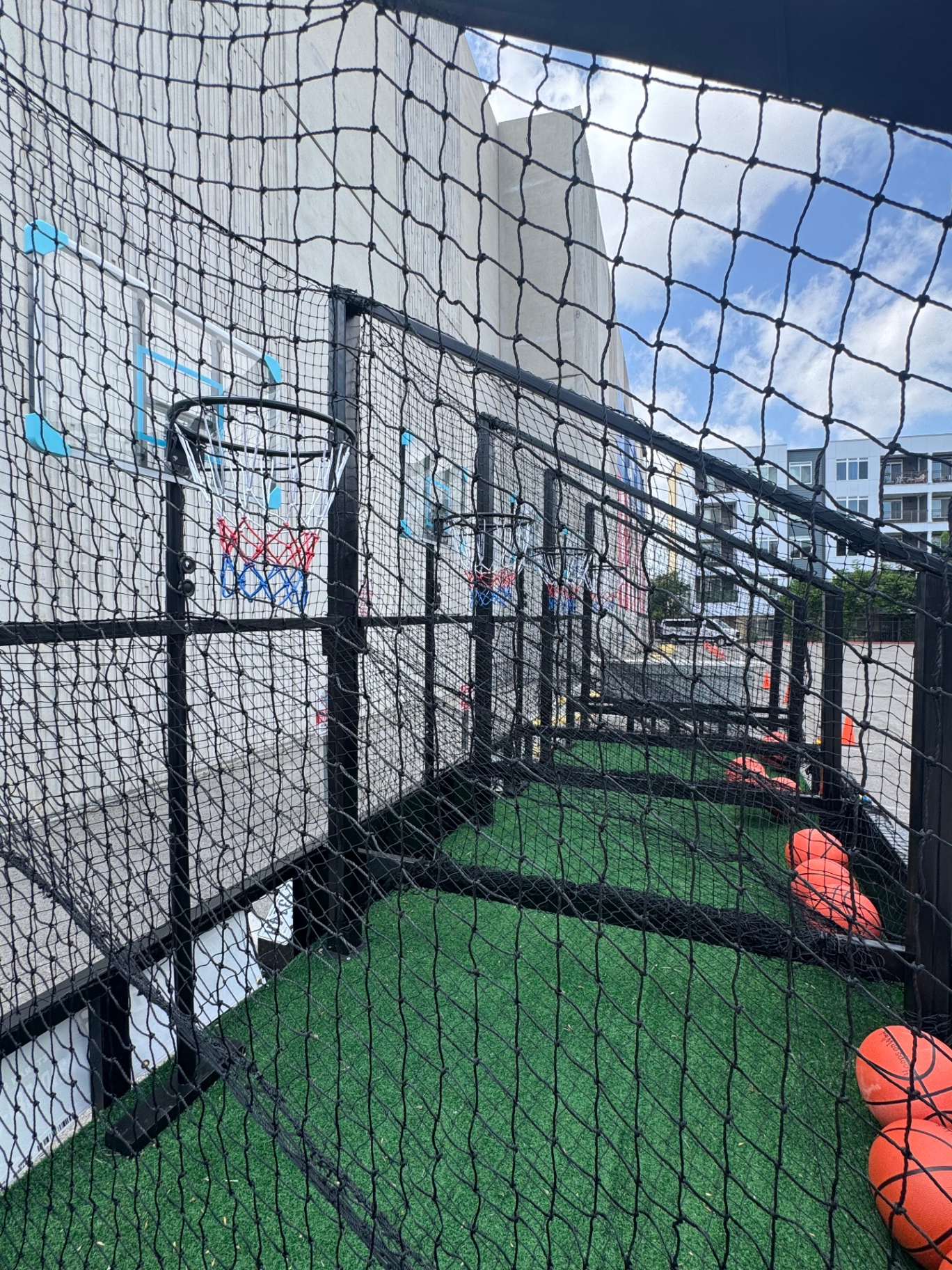 Inside the Hoops on Wheels enclosure, looking down the row of backboards and hoops with basketballs resting on the green turf